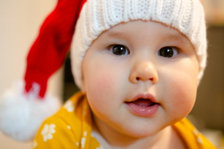 A delightful baby in a cozy Santa hat, radiating holiday joy and innocence. The yellow outfit with cute patterns complements the festive atmosphere, creating a heartwarming scene.の写真素材