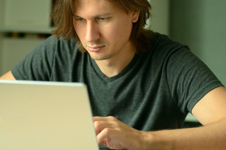 A young man with long hairstyle working on his laptop at home office desk. Focused look at the...