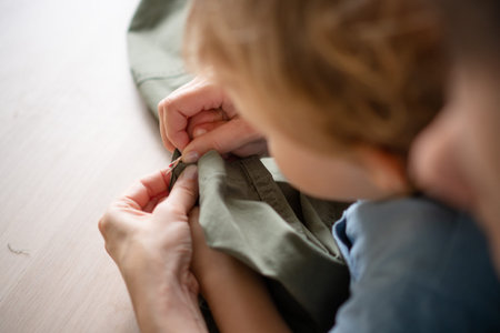 Caring mother dressmaker holding her baby while repairing trousers by unpicking the seam. Top down...