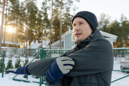 A runner in his forties performs a shoulder stretch outdoors. He warms up on a chilly morning,...