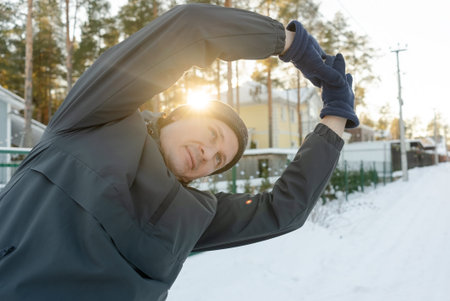A forty-year-old man does side bends with arms raised during sunset. He warms up outdoors in cold...