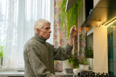 Side view of an older adult stretching arm to reach something on high kitchen shelf. The image...