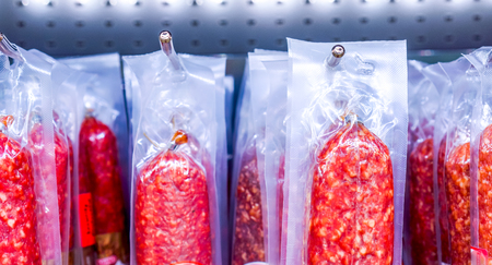 many dry sausages packed in vacuum plastic packaging hanging on metal hooks in supermarket. Selection of dry sausages (background)の写真素材