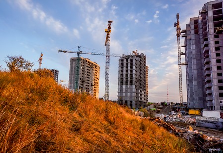 Big future multi-store housing complex with autumn golden yellow grass in sunset sky. Building cranes in front of a multi-storey building under construction which stay one by oneの写真素材