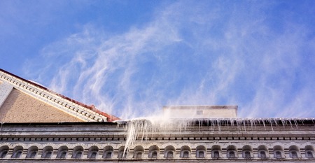 wind blows to roof of old house and snow fly to ground against blue sunny sky with white clouds. Heavy snowfall on a city house roof in the winter time of the yearの写真素材