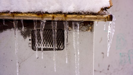 Clear crystal icicles on wooden roof against dark window with grid, white painted wall and snow on roof. Winter hanging icicles on the house roofの写真素材