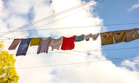 colorful different clothes and big size underclothing hanging on a rope stretched between two brick houses against green tree, sunny blue sky and lines of black wires. Underwear drying on ropeの写真素材