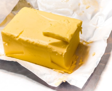 ivory small peace of butter on big butter brick with cracked made by foil package. Ivory block of butter laying on foil wrapping with cracks made by knife. Creamy butter in its unwrapped foil paperの写真素材