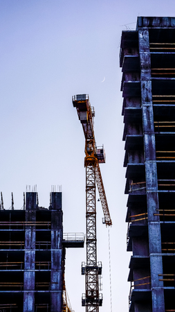 alone crane between buildings against sunset sky. big high crane under a new construction site of high-rise building against blue sunset sky. lonely metal cranes bottom viewの写真素材