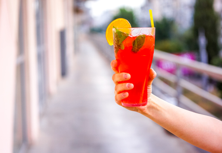 blurry effect on fresh cold red coral fruit watermelon strawberry mojito cocktail with mint, peace of ice, slice of orange and strong cocktail tube in strong man's hand against blurry city background.の写真素材
