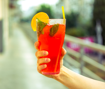 blurry effect on fresh cold red coral fruit watermelon strawberry mojito cocktail with mint, peace of ice, slice of orange and strong cocktail tube in strong man's hand against blurry city background.の写真素材
