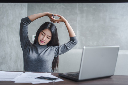 Portrait of young smiling woman wearing suit sitting at home office desk in front of laptop, stretching with enjoyment after the work is done, looking at screen with happy expressionの写真素材