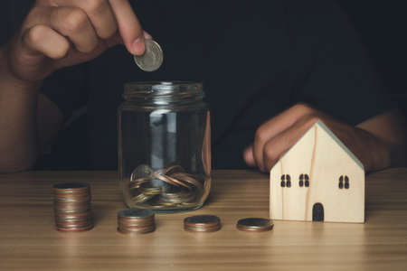Businessman holding coins putting in glass with house wood model and coin stack front, Business,...