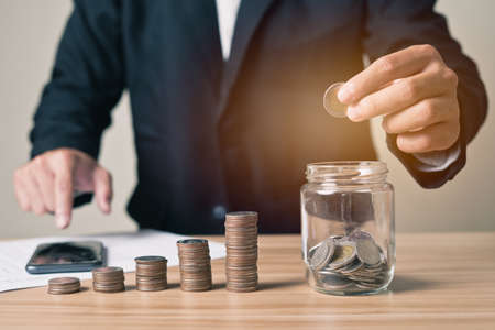Businessman carrying a coin takes it in a glass jar with a pile of coins on the side and  is using a smartphone. Concept investment , finance, businessman, planning, saving money, accounting.の写真素材