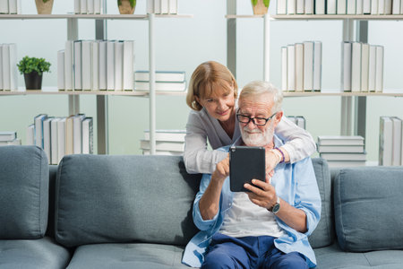 Elderly senior family couple in casual outfit embracing and using smart tablet with smile happy and romantic emotion in living room.の写真素材