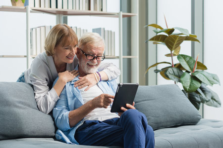 Elderly senior family couple in casual outfit embracing and using smart tablet with smile happy and romantic emotion in living room.の写真素材