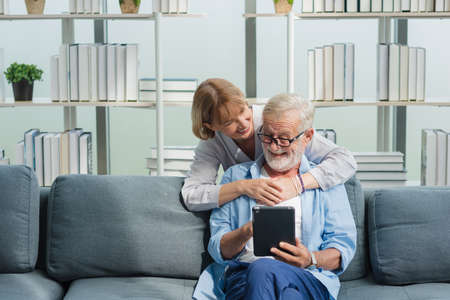 Elderly senior family couple in casual outfit embracing and using smart tablet with smile happy and romantic emotion in living room.の写真素材