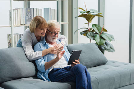 Elderly senior family couple in casual outfit embracing and using smart tablet with smile happy and romantic emotion in living room.の写真素材