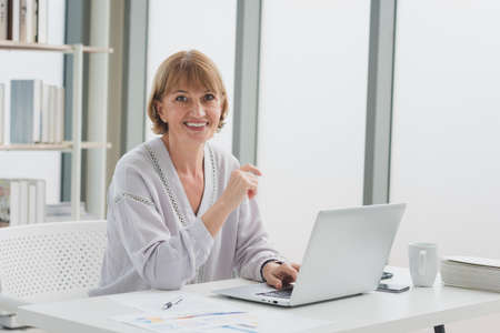 Portrait of elderly senior woman in casual outfit sitting and business working from home with smile and happiness in modern workplace room.の写真素材