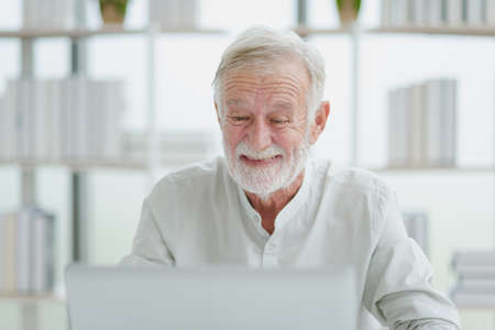 The senior elderly man in white casual outfit work from home sitting and relaxing with happy and smiling emotion in a modern workplace room.の写真素材