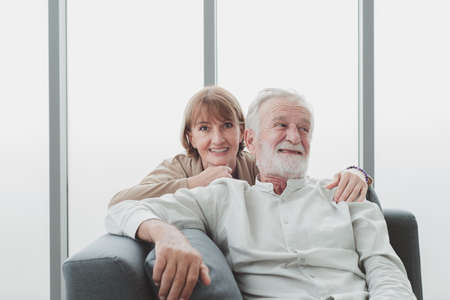 Portrait of senior elderly couple in casual outfit sitting on couch and cuddle together with happy and romantic emotion in living room.の写真素材