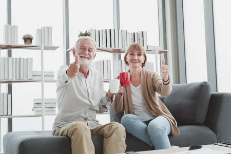 Portrait of senior elderly couple in casual outfit sitting on couch and drink a cup of coffee together with happy and romantic emotion in living room.の写真素材