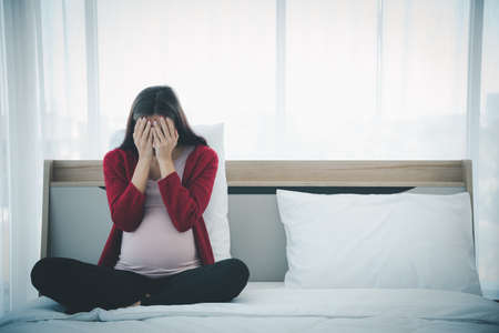 Pregnant woman in casual home outfit sitting on bed and feeling upset and sad from family conflict...
