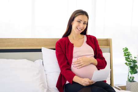 Portrait of pregnant woman in casual home outfit sitting on bed and looking at her ultrasound image of her child in her belly in modern bedroom.の写真素材