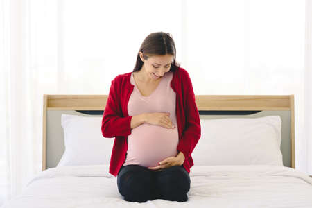 Portrait of pregnant woman in casual home outfit sitting on bed and holding child in her belly in modern bedroom.の写真素材