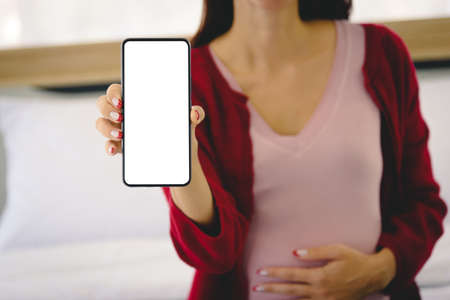 Pregnant woman sitting on bed and holding smart phone with white background for editing in modern bedroom.の写真素材