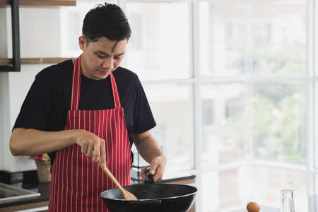 Young Asian man in casual outfit wearing apron cooking food for breakfast in modern interior kitchen room.の写真素材