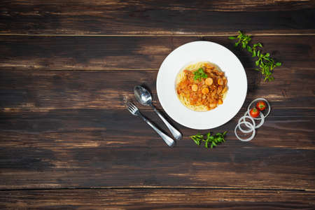 Spaghetti with sausages and organic tomato sauce in plate on top with parsley and beside with spoon, fork, tomato, onion slices and green leaf all in dark wood background.の写真素材