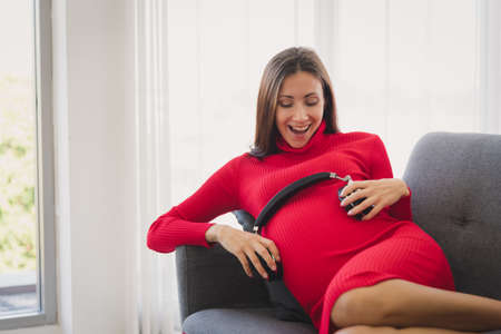 Pregnant woman in red sweater dress lay down on couch and listening to the music and put on the...