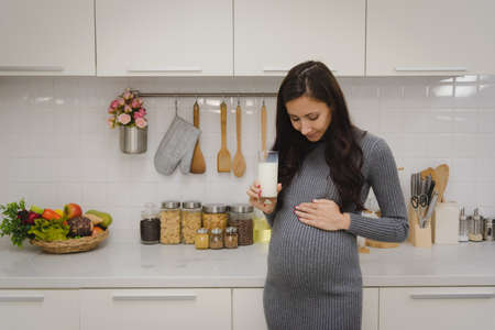 Pregnant woman in grey sweater dress holding her belly and drink a glass of milk in modern kitchen room.の写真素材