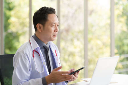 Asian Doctor man in white professional lab coat uniform sitting and using laptop in modern workplace office.の写真素材