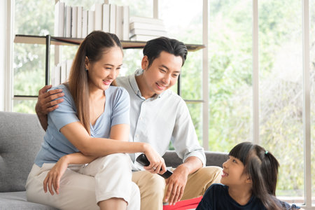 Happy mother father and daughter family sitting on couch and watching television together relaxing at home.の写真素材