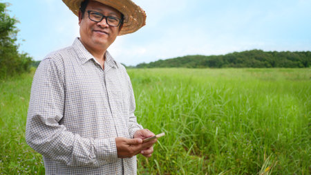 A farmer is using a mobile phone standing in a field against a green field backdrop. The idea of...