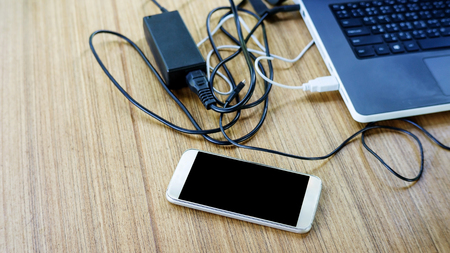 Close up of a smartphone, laptop computer and many cables on a wooden table.の写真素材