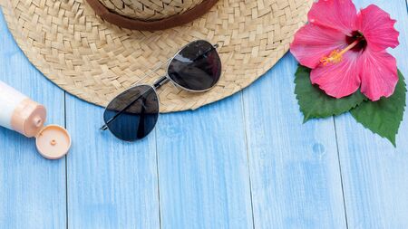 Hat, sunglasses, sunblock and Chinese rose flower on a blue wooden table.