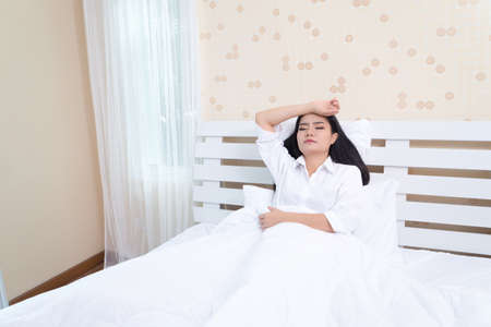 Beautiful girl in the morning, lying on a white bed in a white shirt with headache / insomnia / migraine / stressの写真素材