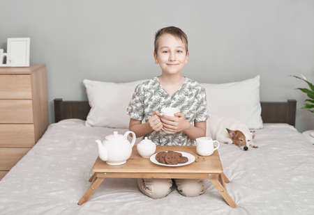 Happy boy sitting on bed and drink tea. Empty space for text. Friendship concept. Cute little boy with funny dog on bed at home. Two friends boy and dog lying together on bedの写真素材