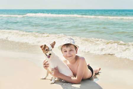 Child boy with dog jack russel on beach. Best friends rest on vacation, play in sand against sea. Tourism and vacation on ocean. Family vacation in summer. Vacation at sea with dog. Summer travelの写真素材