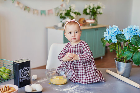 Cute baby girl in a red checkered dress sits at a table in the kitchen and kneads dough.