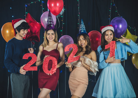 Happy young group of asian friends celebrating Christmas or New Year Party, wearing party hats and holding red shiny paper numeral 2021, having bright colorful balloons with light decoration backgroundの写真素材