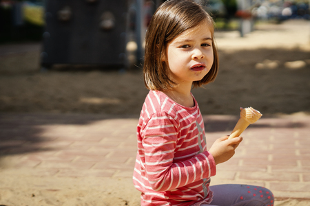 beautiful little girl eats ice cream sitting on the playground in summerの写真素材