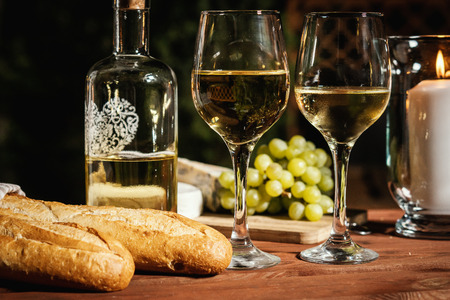 romantic dinner with two glasses of wine, a baguette and snacks on an old wooden table on a summer eveningの写真素材