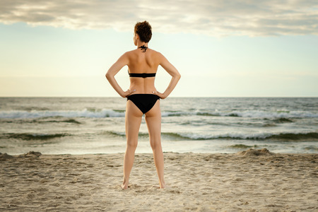 beautiful woman in a black swimsuit is looking at the sea on the beach at sunset