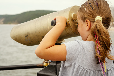 little girl in a dress is looking through a lunete on the sea in summerの写真素材