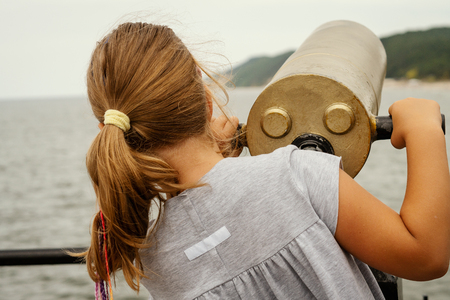 little girl in a dress is looking through a lunete on the sea in summerの写真素材