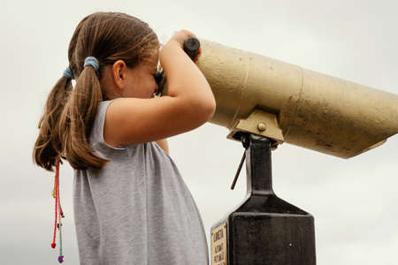 little girl in a dress is looking through a lunete on the sea in summerの写真素材